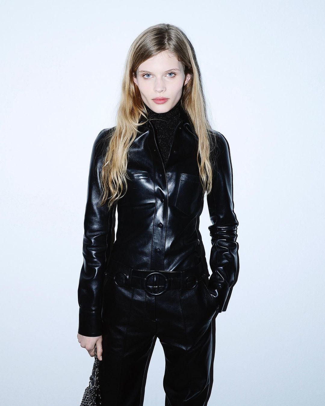 Portrait of a blonde woman in a shiny black jumpsuit against a bright white backdrop in a fashion setting, looking directly at the camera.