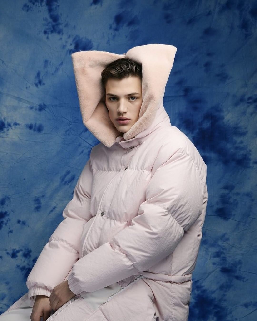Fashion shot of a male model in a pink puffer jacket with a fur-lined hood, posing against a blue tie-dye backdrop in a studio setting.