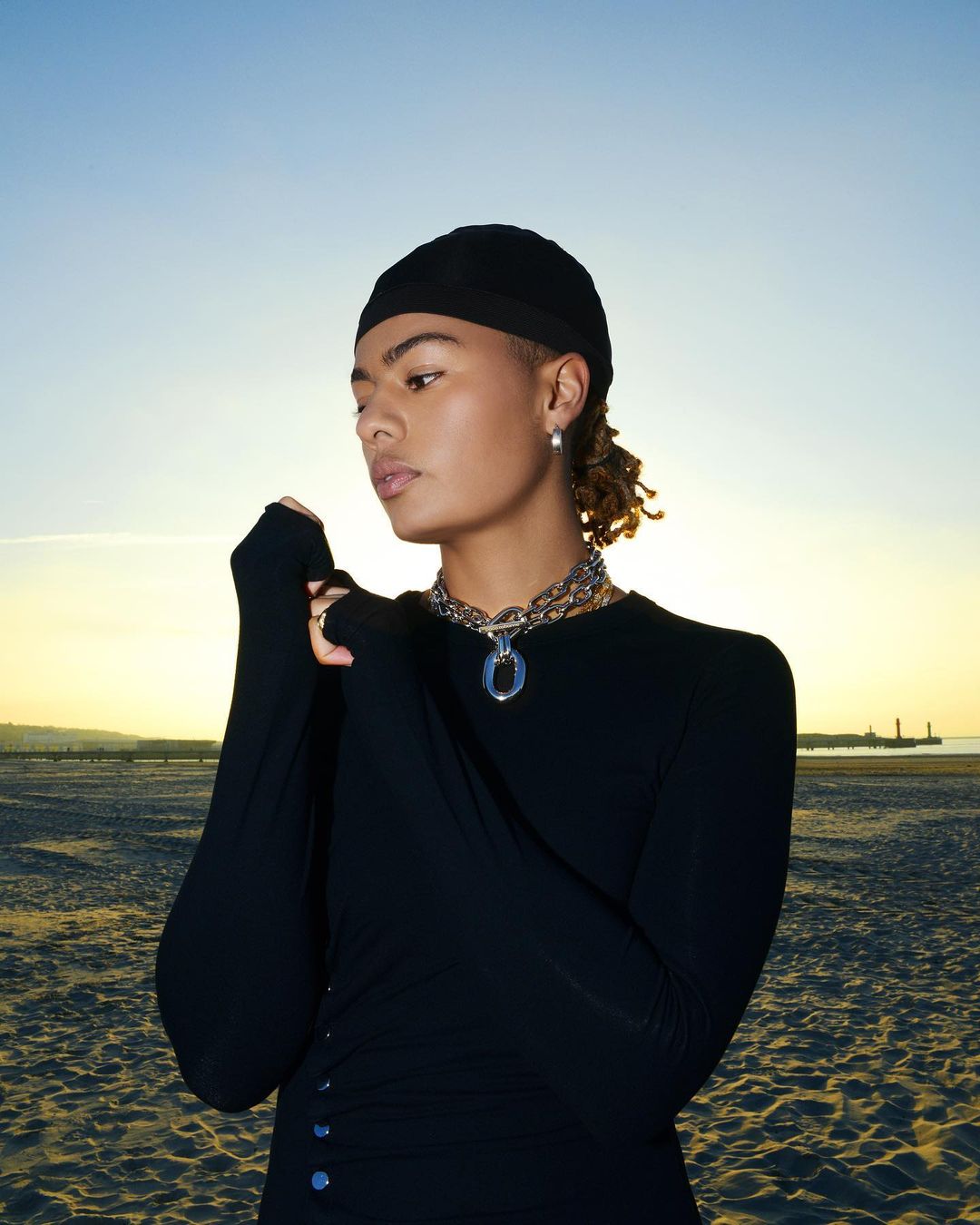 Portrait of a mixed race young adult wearing a black shirt and cap, posing on a beach at sunset with the sky visible.
