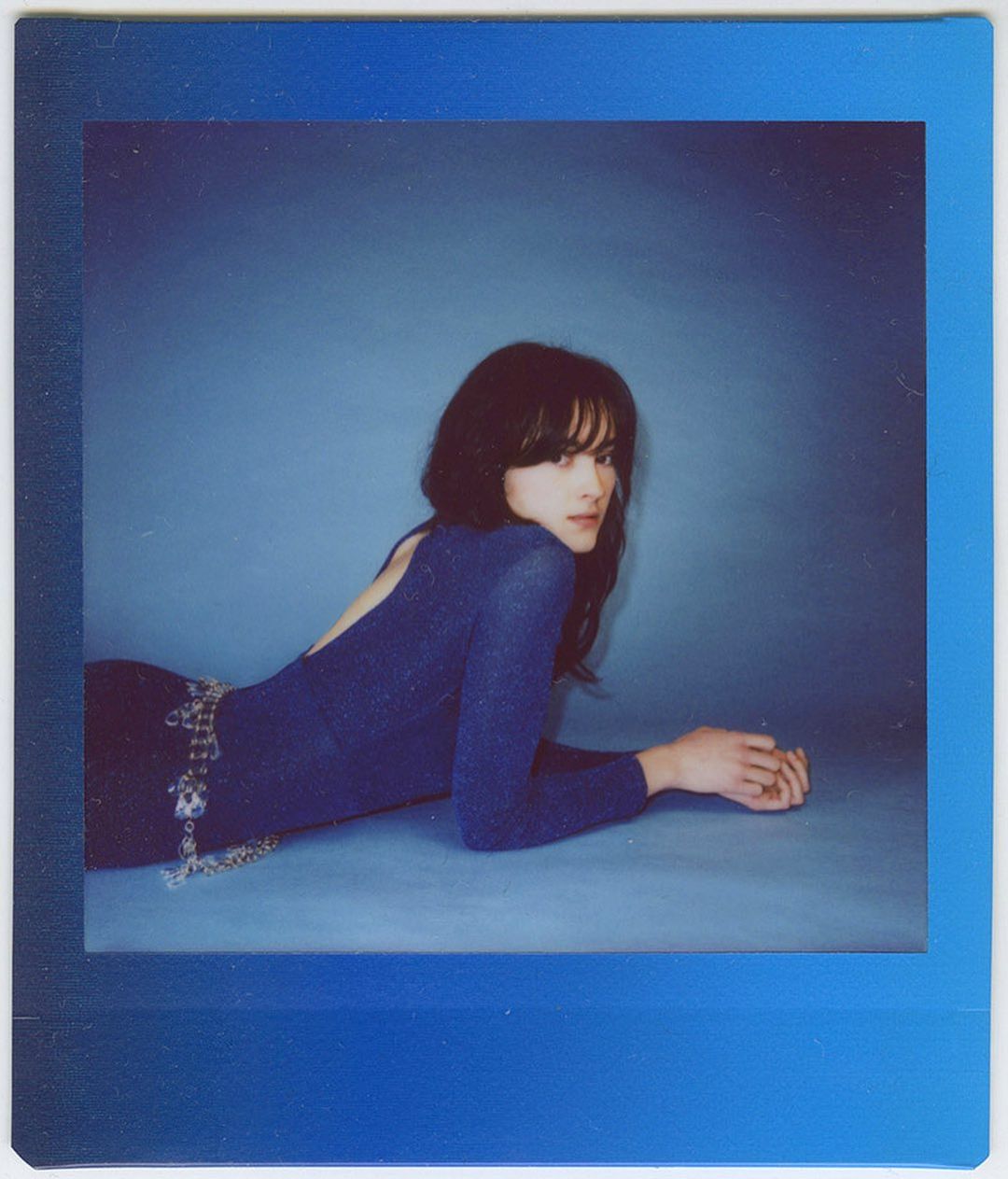 Brunette woman with bangs in a blue bodysuit and silver belt poses in front of a blue backdrop in a polaroid-style frame.
