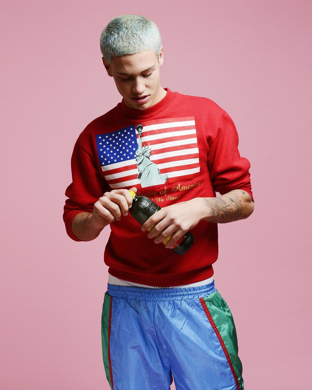 Young man in a red sweatshirt with a US flag graphic opens a bottle of juice against a pink backdrop, creating a vibrant, casual portrait.