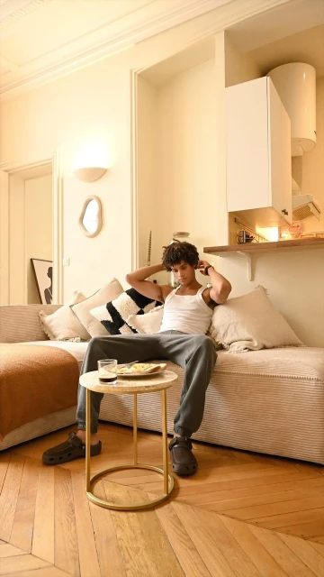 Man relaxes on tan sofa in casual wear. Table with plate and drink in bright living room with parquet floor and decorative wall elements.