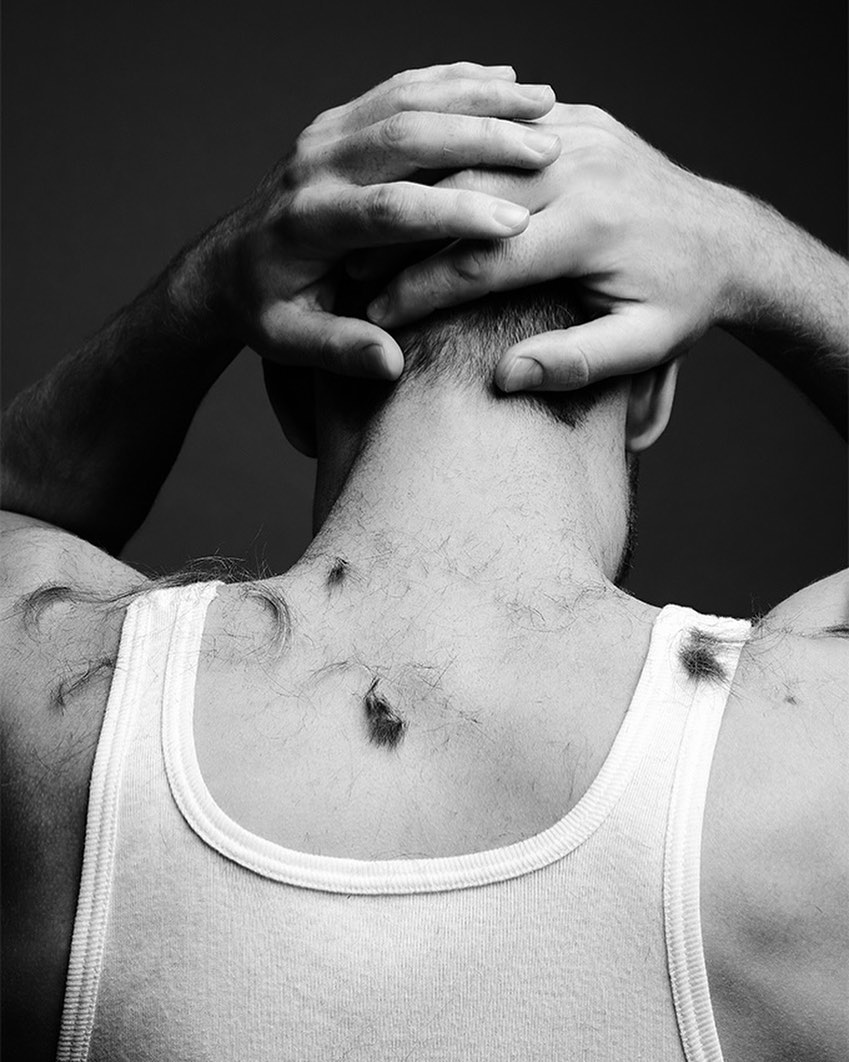 Back view of an adult man in a white tank top with his hands clasped behind his head and hair clippings on his shoulders.