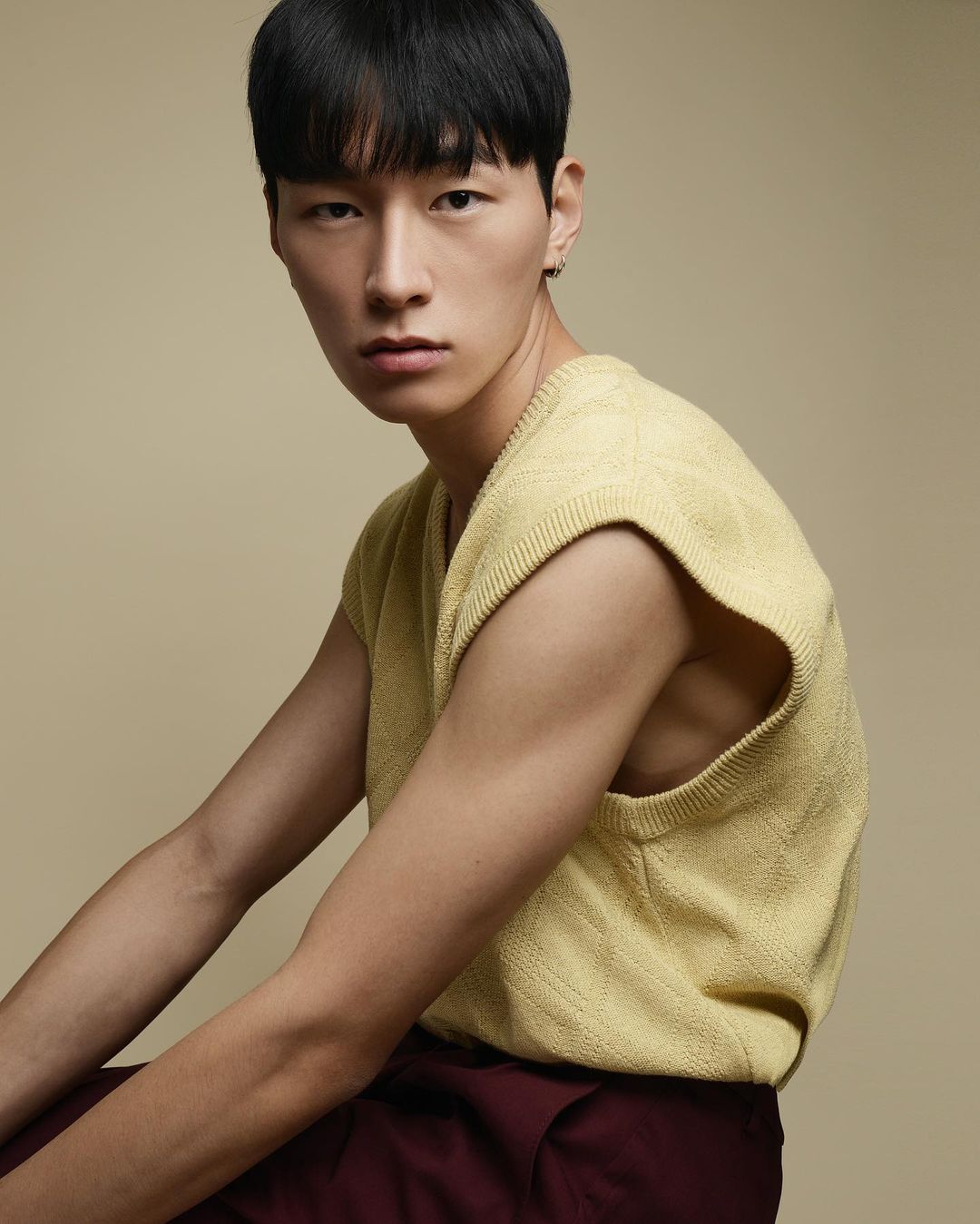Portrait of a young adult Asian male wearing a yellow knit vest and maroon pants, posing in a studio with a neutral background.