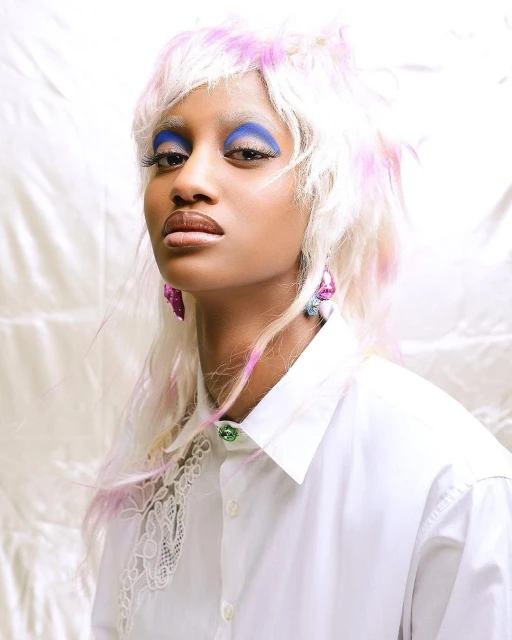 Portrait of a young woman with bright blue eye makeup, white and pink wig, wearing a white button-down shirt in a light-filled studio.