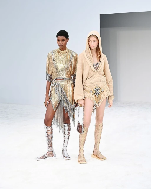 Two models on a white runway showcase gold and beige embellished outfits with gladiator sandals, high fashion in a minimalist setting.