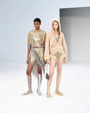 Two models on a white runway showcase gold and beige embellished outfits with gladiator sandals, high fashion in a minimalist setting.