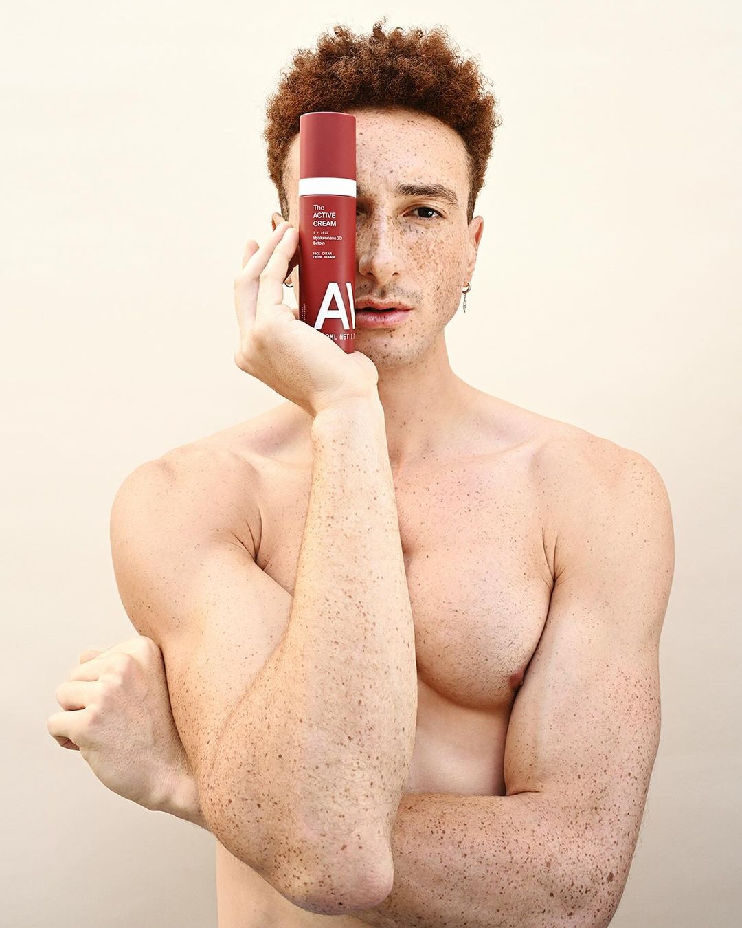 Man poses holding red cosmetic bottle in front of face. Red hair and light skin with freckles against a light background.