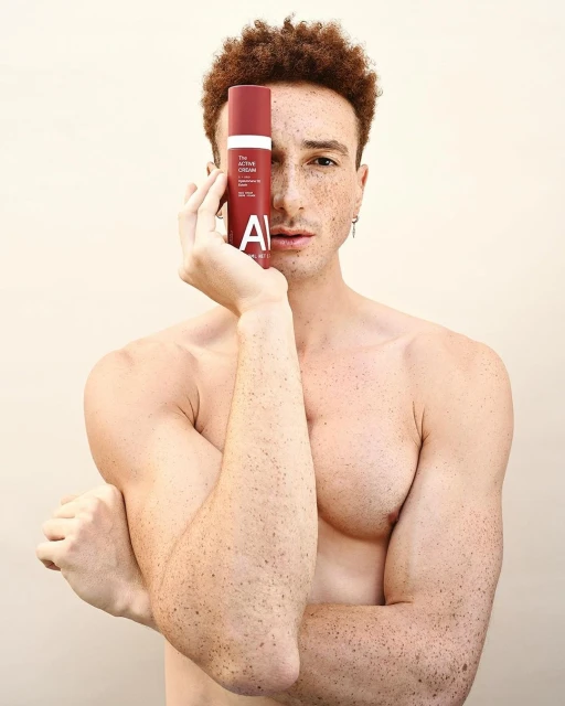 Man poses holding red cosmetic bottle in front of face. Red hair and light skin with freckles against a light background.