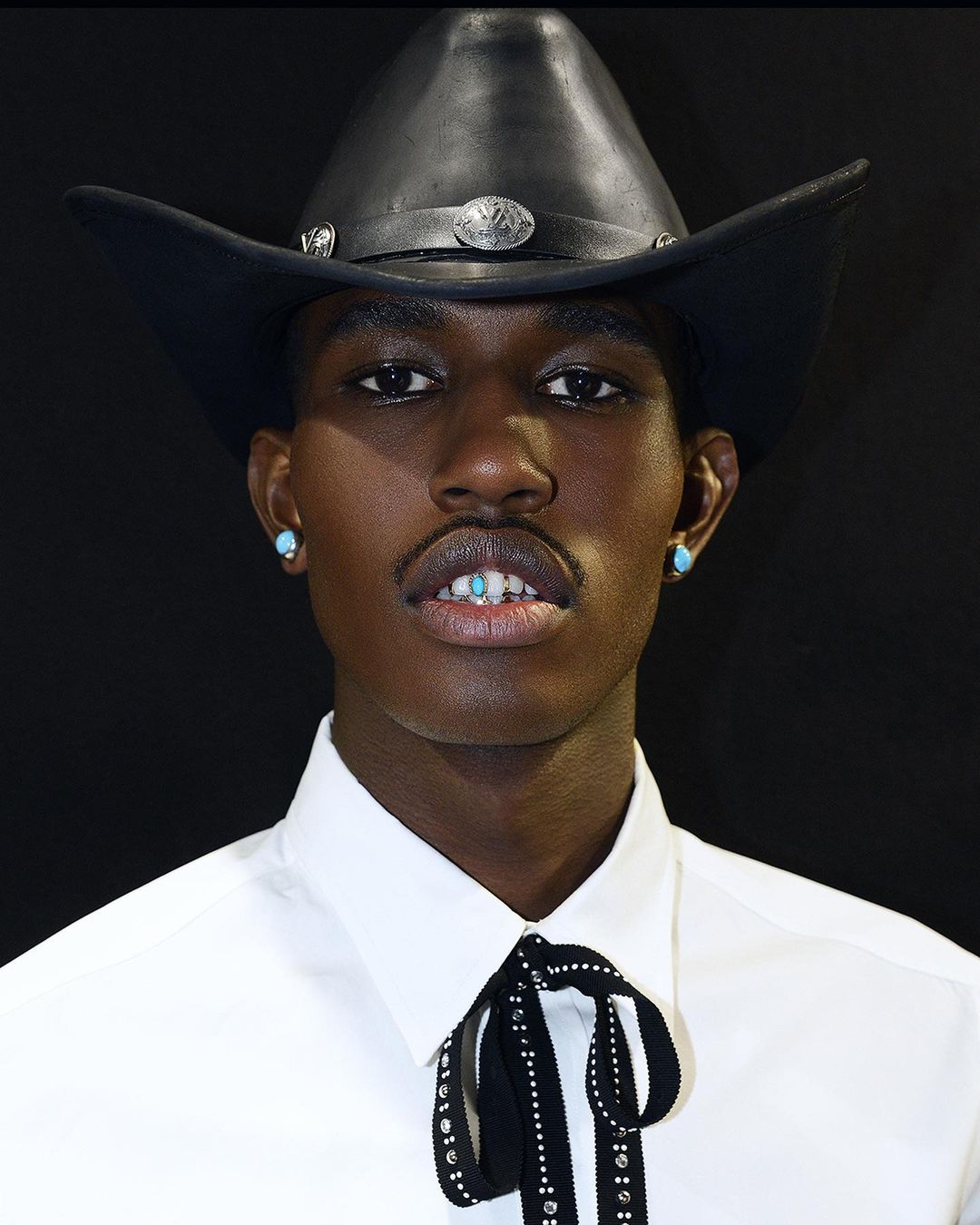 Close-up of a Black adult male wearing a cowboy hat, white shirt, and turquoise earrings against a dark background.
