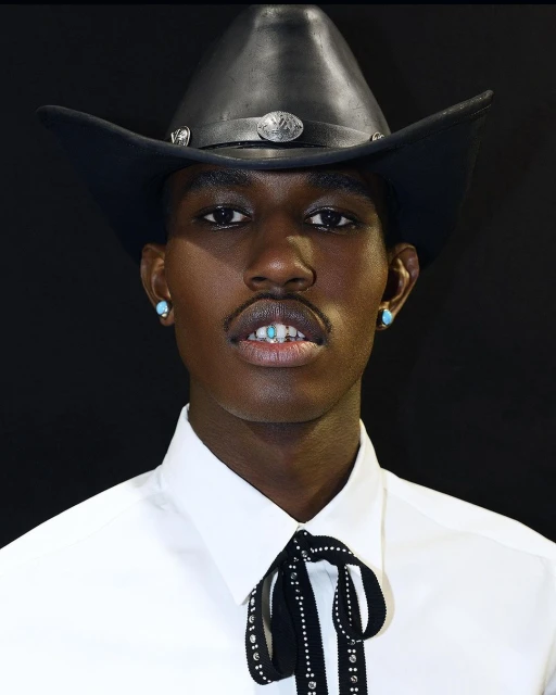 Close-up of a Black adult male wearing a cowboy hat, white shirt, and turquoise earrings against a dark background.