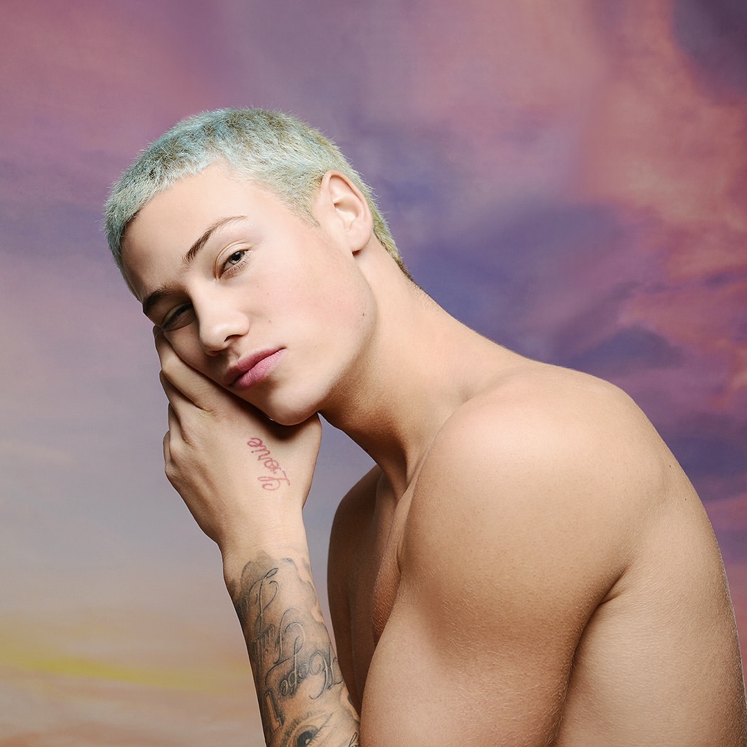 Portrait of a young adult with blue-tinted hair, fair skin, and a tattoo on hand, posing against a blurred sky background.