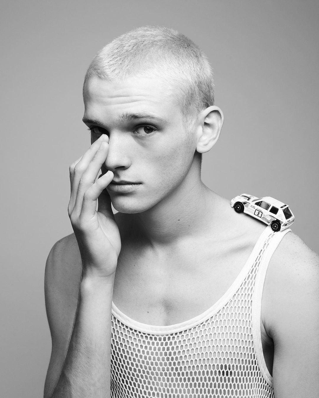 Black and white portrait of a light-skinned young adult with short hair, wearing a mesh shirt with a toy car on his shoulder.