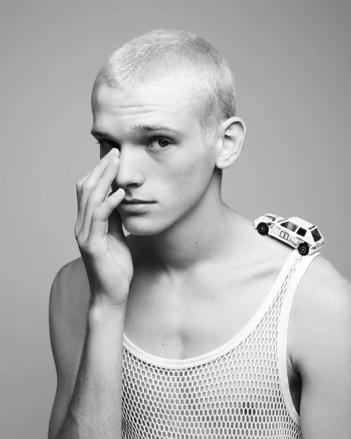 Black and white portrait of a light-skinned young adult with short hair, wearing a mesh shirt with a toy car on his shoulder.