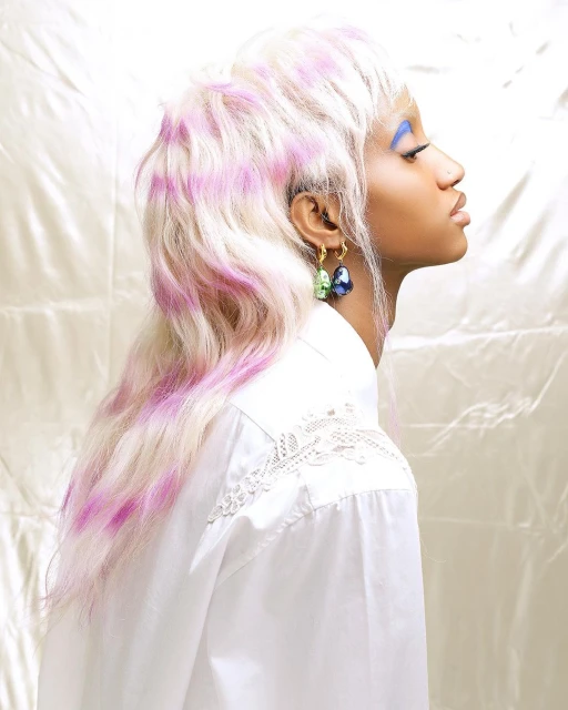 Side profile of a woman with dyed white and pink hair, blue eyeshadow, wearing a white shirt and statement earrings against a shiny backdrop.