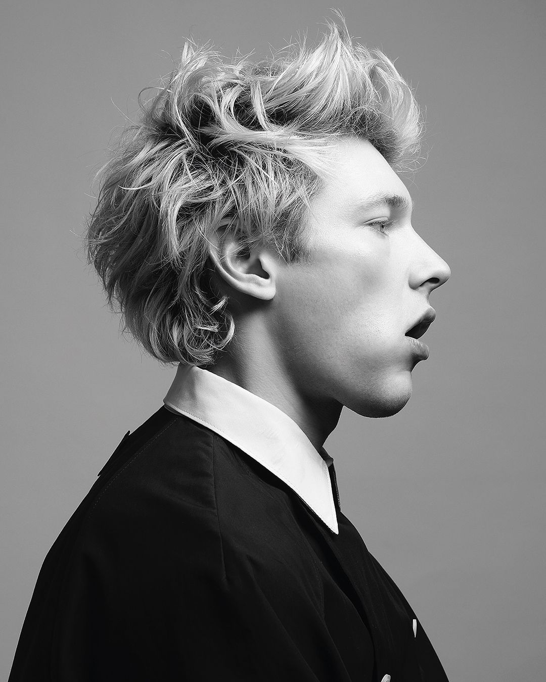 Black and white portrait of a young adult male with tousled hair, wearing a collared shirt and jacket, captured in profile against a grey backdrop.