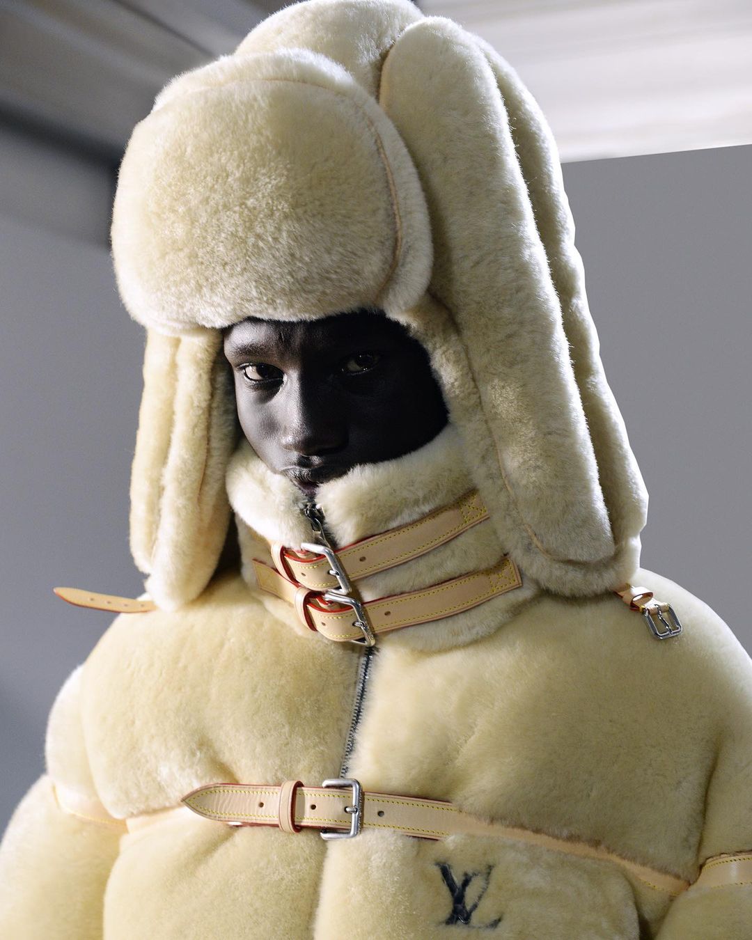 Fashion portrait of a dark-skinned adult male wearing a light-colored fur trapper hat and a belted down coat with brand logo.