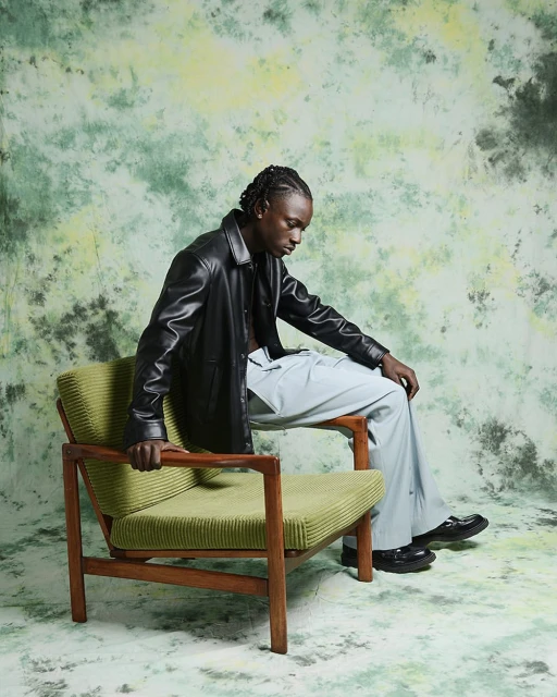 A young Black adult male in a black leather jacket and light blue pants sits in a green armchair against a mottled green backdrop.