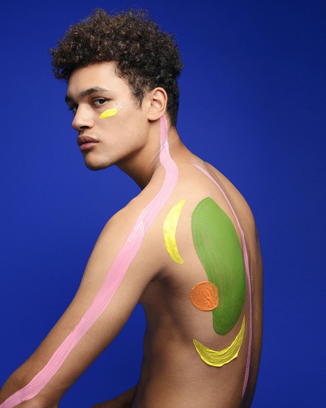 Shirtless adult male poses against a blue backdrop with colorful body paint designs across the torso and face in a studio setting.
