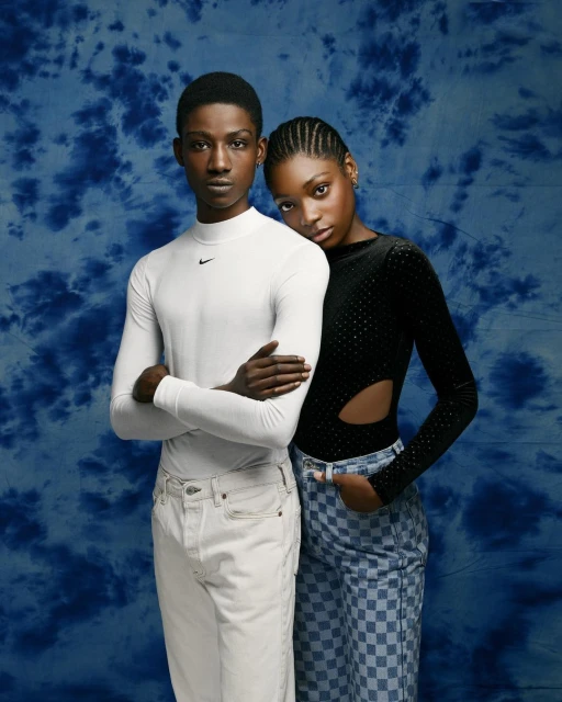 Two Black adults, one standing and one leaning on their shoulder, both wearing jeans against a blue and white mottled background.