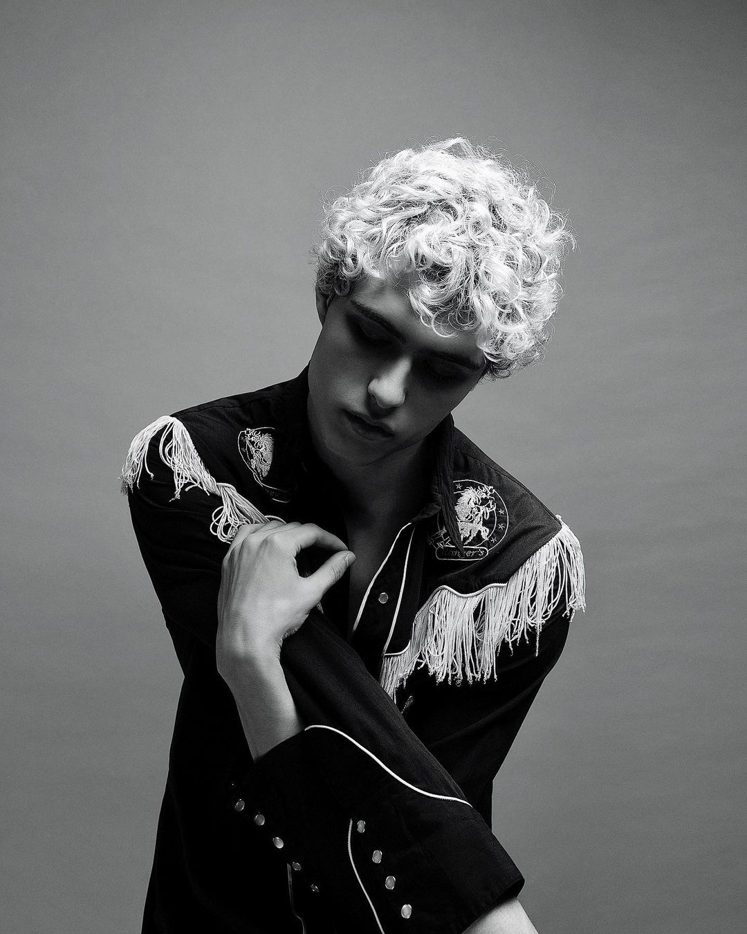 Monochrome portrait of a young adult male with curly hair, wearing a fringed Western-style shirt against a neutral grey backdrop.