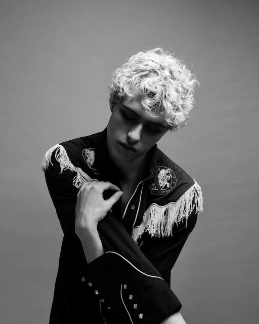 Monochrome portrait of a young adult male with curly hair, wearing a fringed Western-style shirt against a neutral grey backdrop.