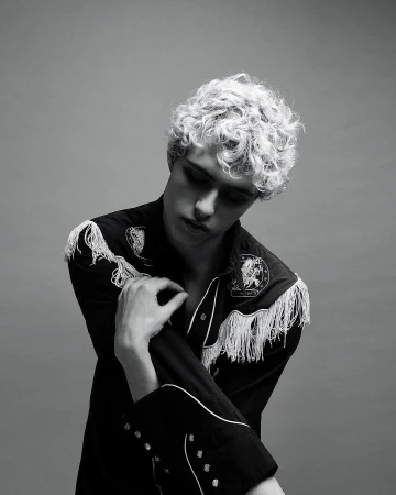 Monochrome portrait of a young adult male with curly hair, wearing a fringed Western-style shirt against a neutral grey backdrop.