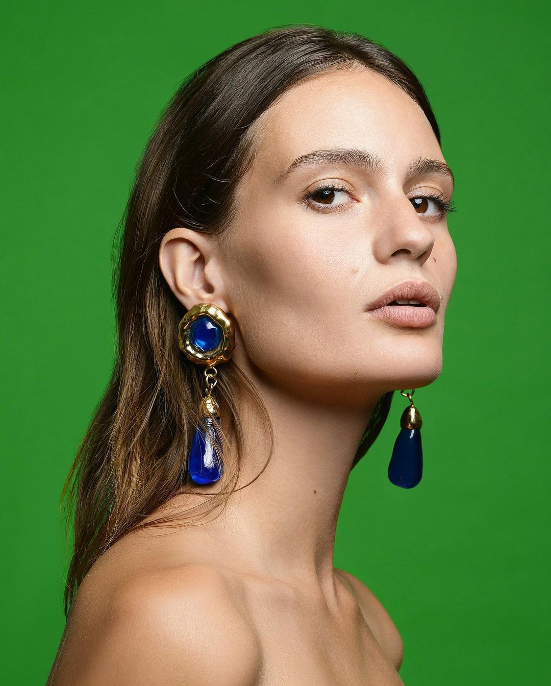 Portrait of a young adult woman against green background, wearing dangling blue and gold earrings. Fashion and beauty photography.