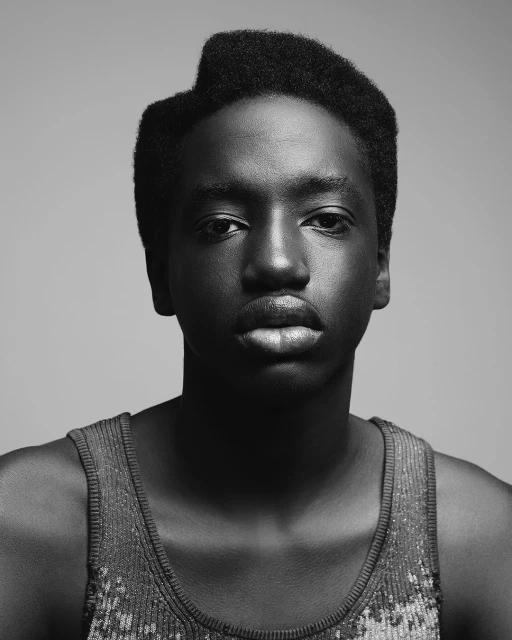 Close-up portrait of an African American male with a distinctive hairstyle wearing a sequined tank top against a neutral gray backdrop.