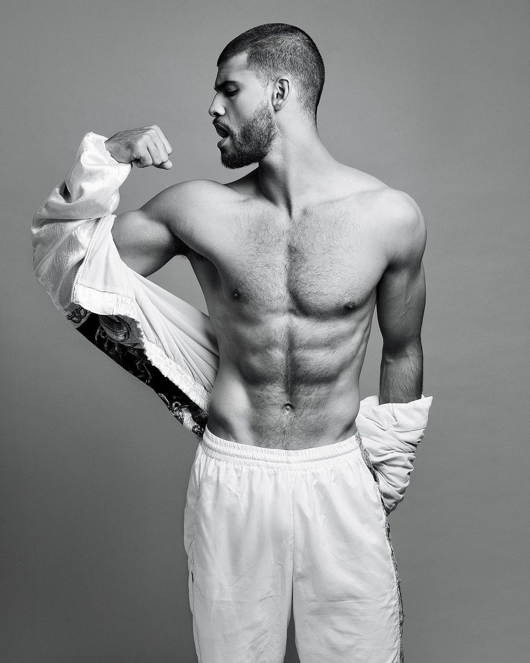 Black and white photo of a muscular, bearded man flexing his bicep while wearing a jacket off one shoulder and athletic pants against gray background.