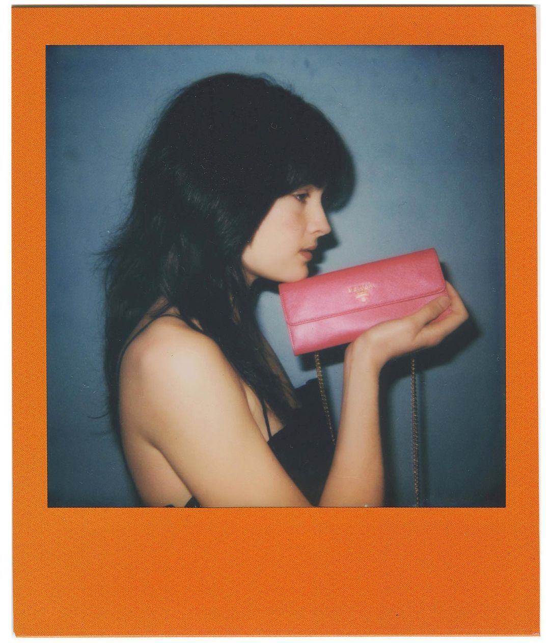 Woman in profile holding a pink Prada wallet with a chain strap against a blue background in an orange Polaroid frame.