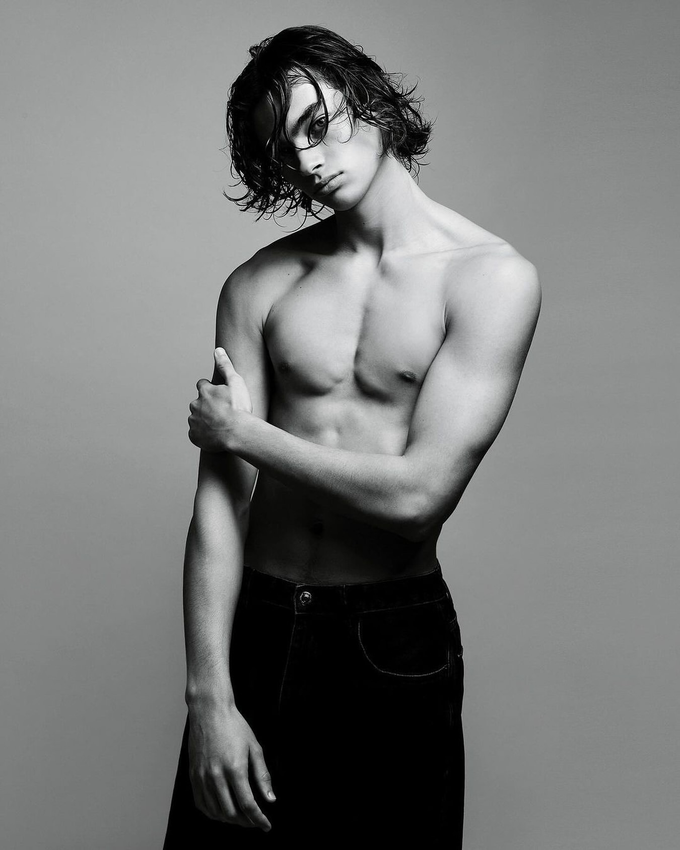 Black-and-white portrait of a shirtless young adult male model with wet hair, striking a pose in dark trousers against a gray backdrop.