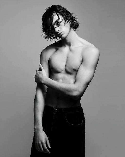 Black-and-white portrait of a shirtless young adult male model with wet hair, striking a pose in dark trousers against a gray backdrop.