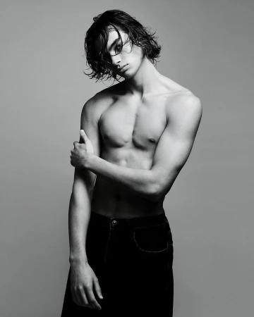 Black-and-white portrait of a shirtless young adult male model with wet hair, striking a pose in dark trousers against a gray backdrop.