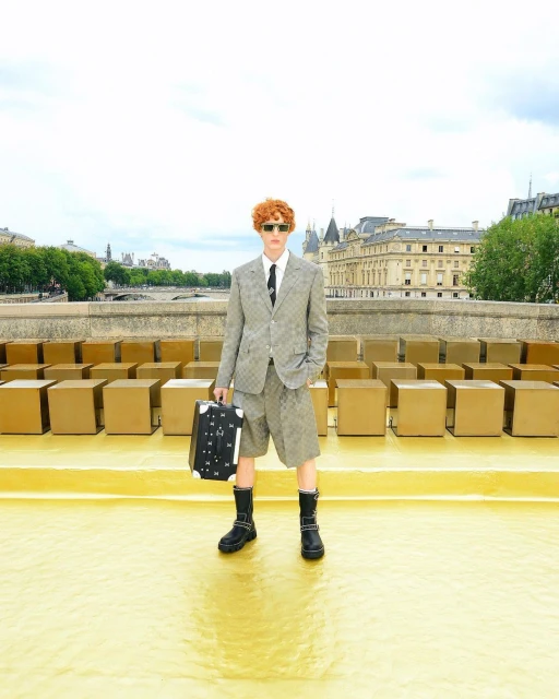 Man in patterned suit and boots standing on gold platform with patterned suitcase and background of tan boxes and European buildings.