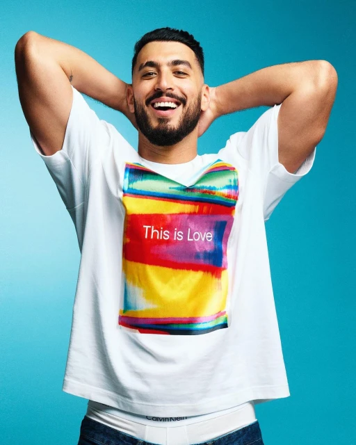 Smiling man poses in white t-shirt with colorful graphic print on blue background, looking directly at camera in medium shot.