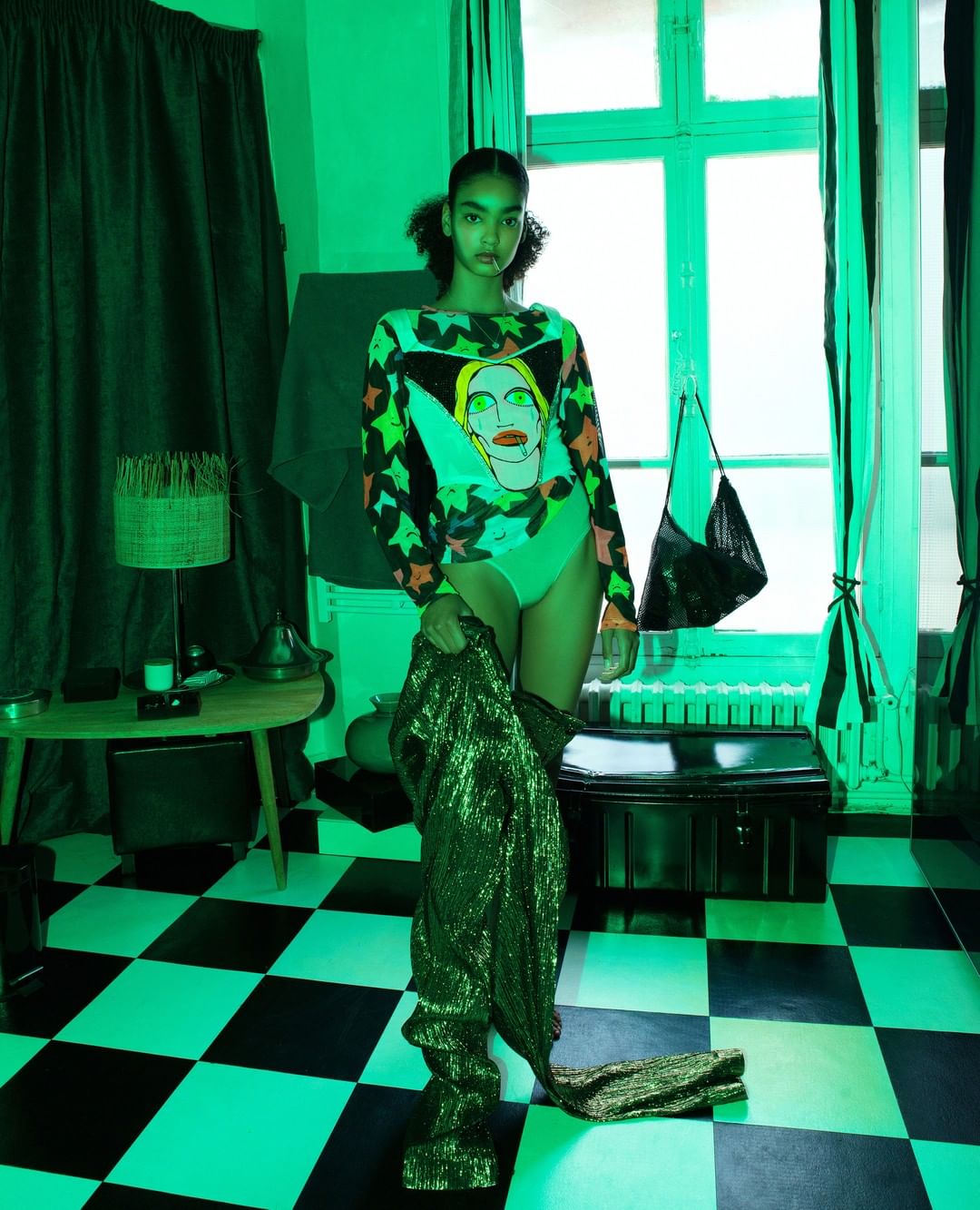 Young woman poses in green-toned room, wearing a graphic long-sleeve shirt with a face, leotard, and shiny green thigh-high boots.
