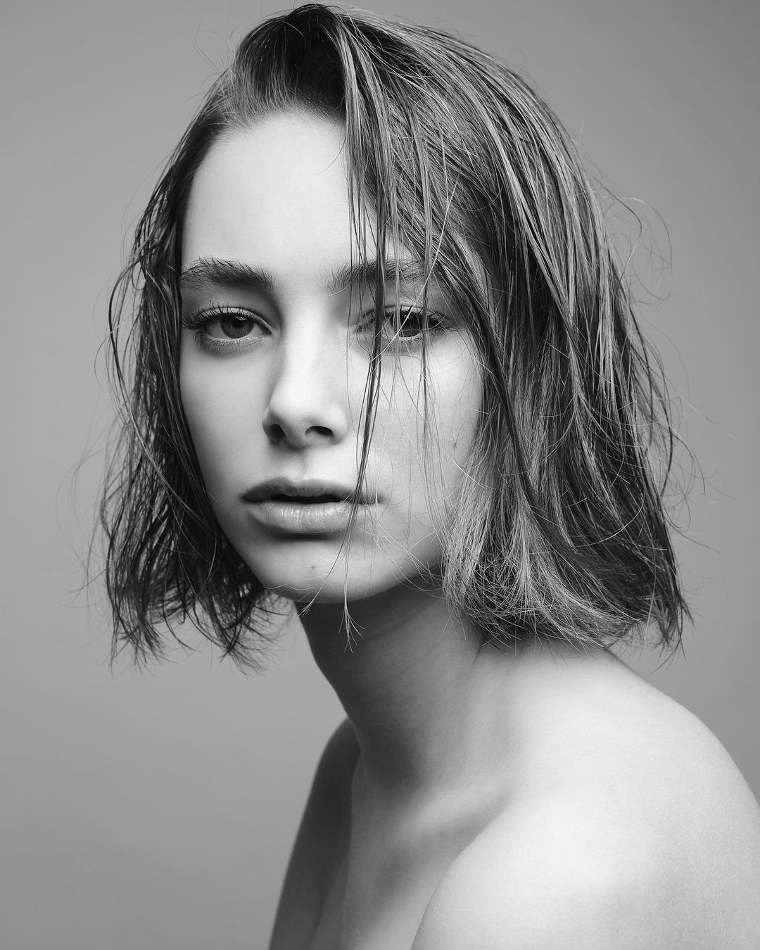 Monochrome portrait of a young woman with damp, tousled hair and a direct gaze against a neutral background.