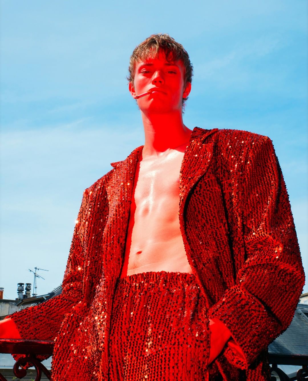 A young adult male in a red sequined suit stands on a balcony under a blue sky, holding an object in his mouth, posing for a fashion shot.