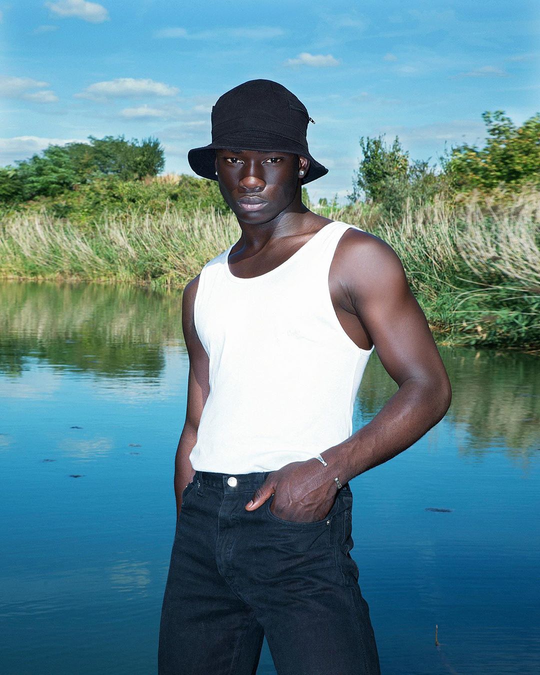 Portrait of a dark-skinned man in a white tank top and black bucket hat stands in front of a body of water on a bright day.
