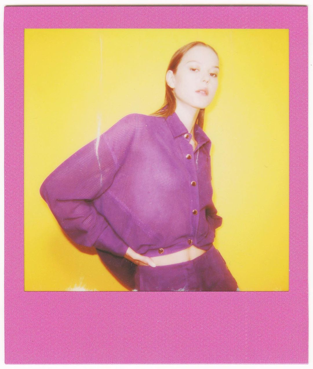 Eye-level studio shot of a young woman posing in purple knitwear against a bright yellow background, framed by a pink border.