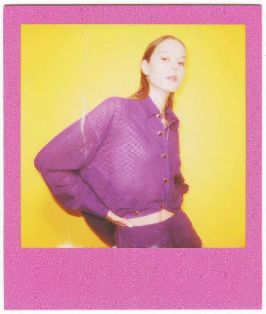 Eye-level studio shot of a young woman posing in purple knitwear against a bright yellow background, framed by a pink border.