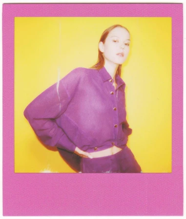 Eye-level studio shot of a young woman posing in purple knitwear against a bright yellow background, framed by a pink border.
