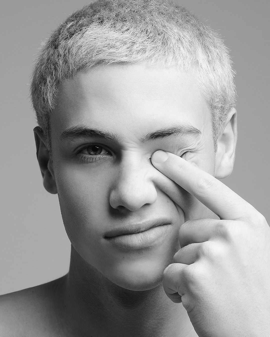 Close-up black and white portrait of a young adult male with fair skin and short light hair, touching his face, on gray background.