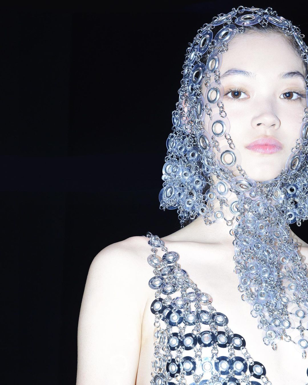 Asian woman wearing a unique metal headpiece and matching metal top in a studio setting, posing against a dark backdrop. Fashion shoot.
