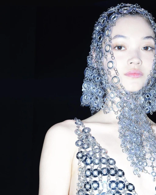 Asian woman wearing a unique metal headpiece and matching metal top in a studio setting, posing against a dark backdrop. Fashion shoot.