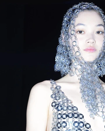 Asian woman wearing a unique metal headpiece and matching metal top in a studio setting, posing against a dark backdrop. Fashion shoot.