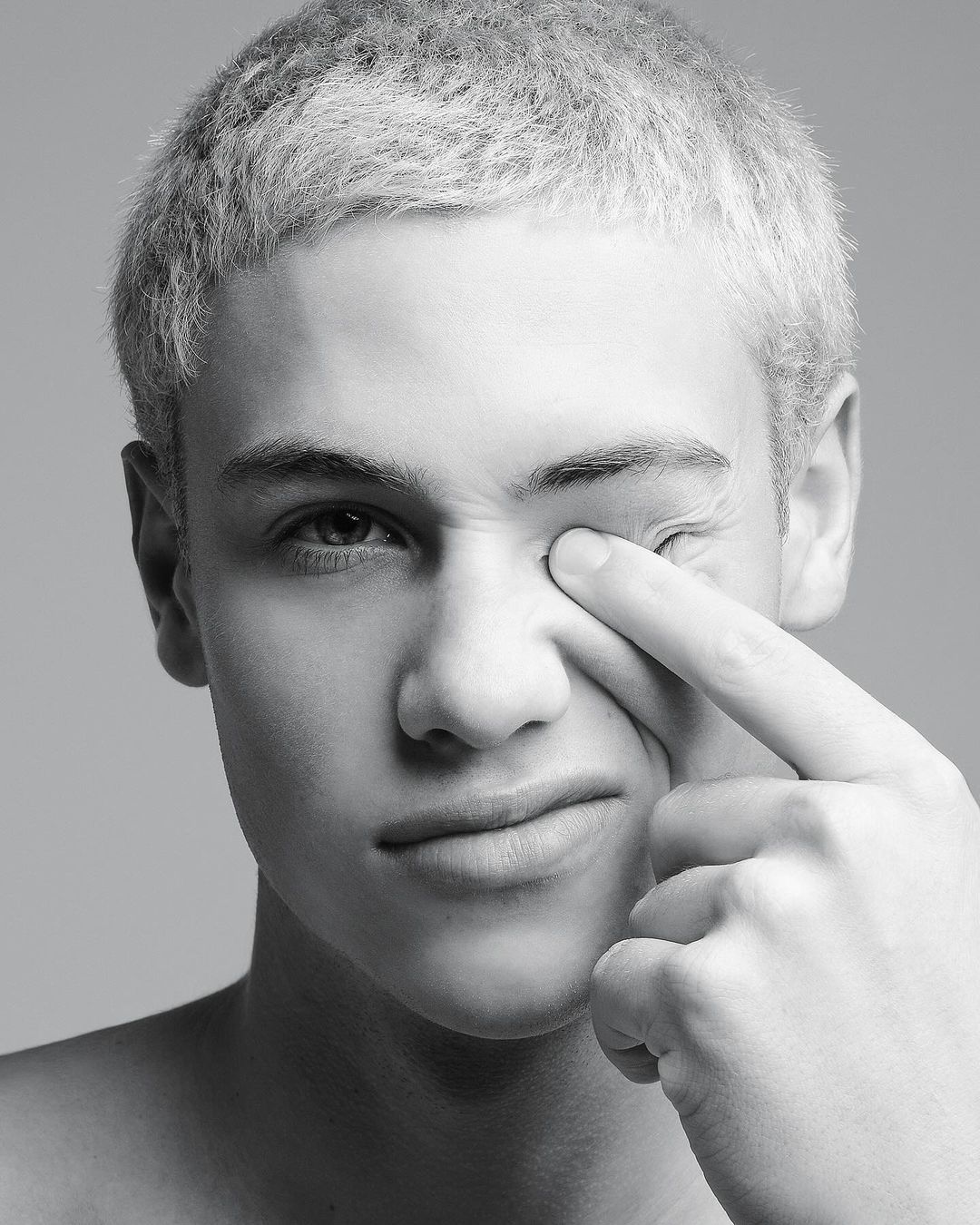 Close-up black and white portrait of a young adult male with short blond hair touching his eye, set against a neutral gray background.