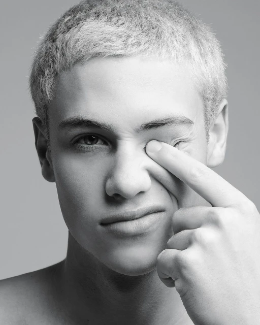 Close-up black and white portrait of a young adult male with short blond hair touching his eye, set against a neutral gray background.
