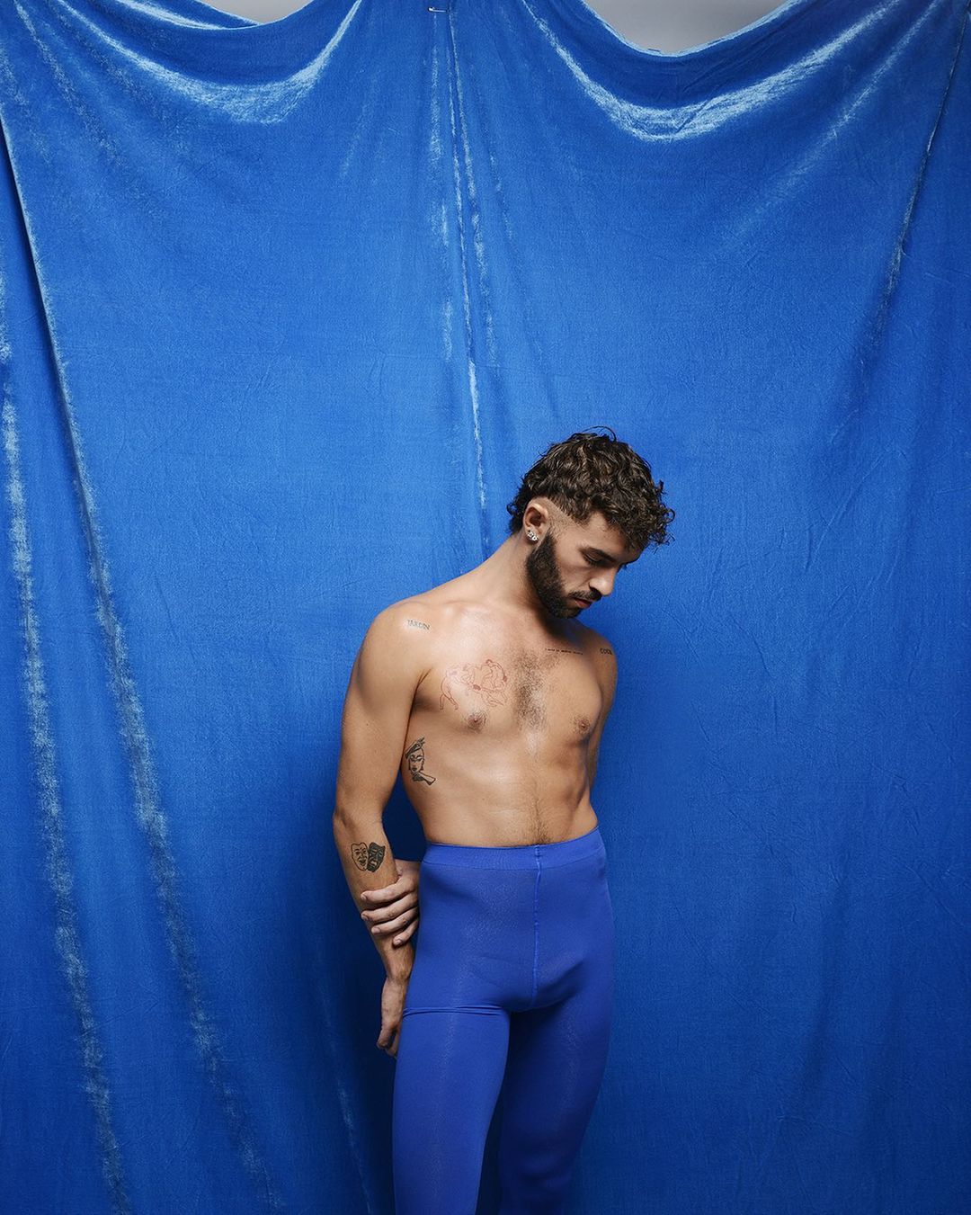 Shirtless male in blue leggings poses against a draped blue velvet backdrop, looking down with curly brown hair and beard in a studio setting.